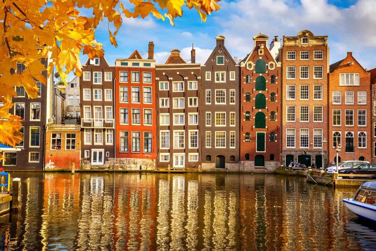 Autumn leaves frame a tranquil Amsterdam canal reflecting the charming facades of historic Dutch buildings in a palette of warm colors, with a moored boat completing the picturesque scene