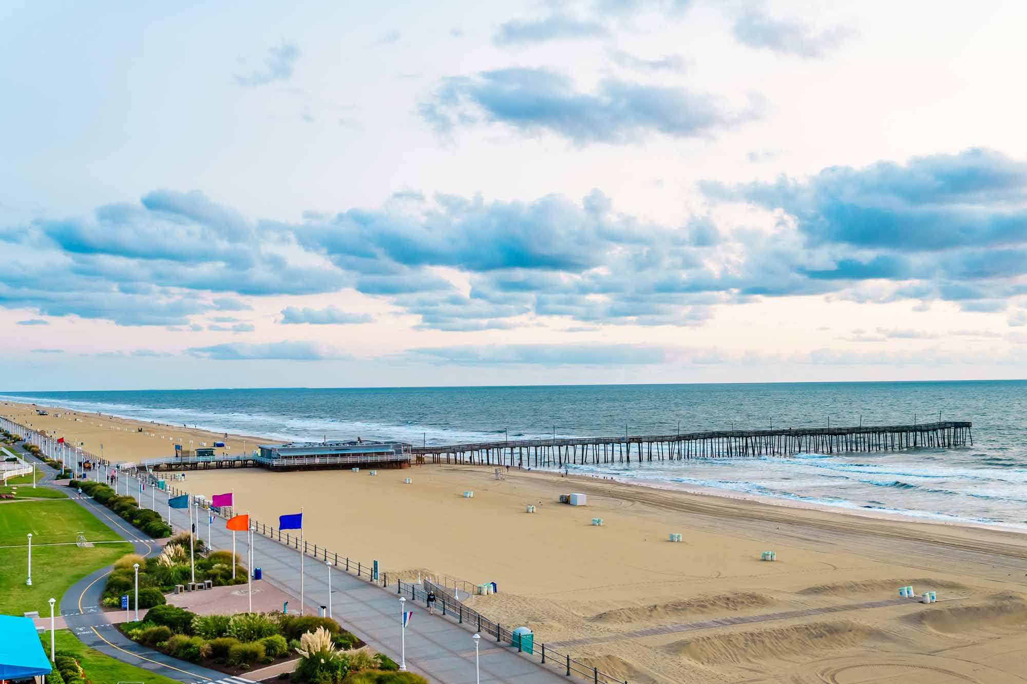 A picturesque view of Virginia Beach showing a wide sandy beach, a long pier, and a promenade under a cloudy sky