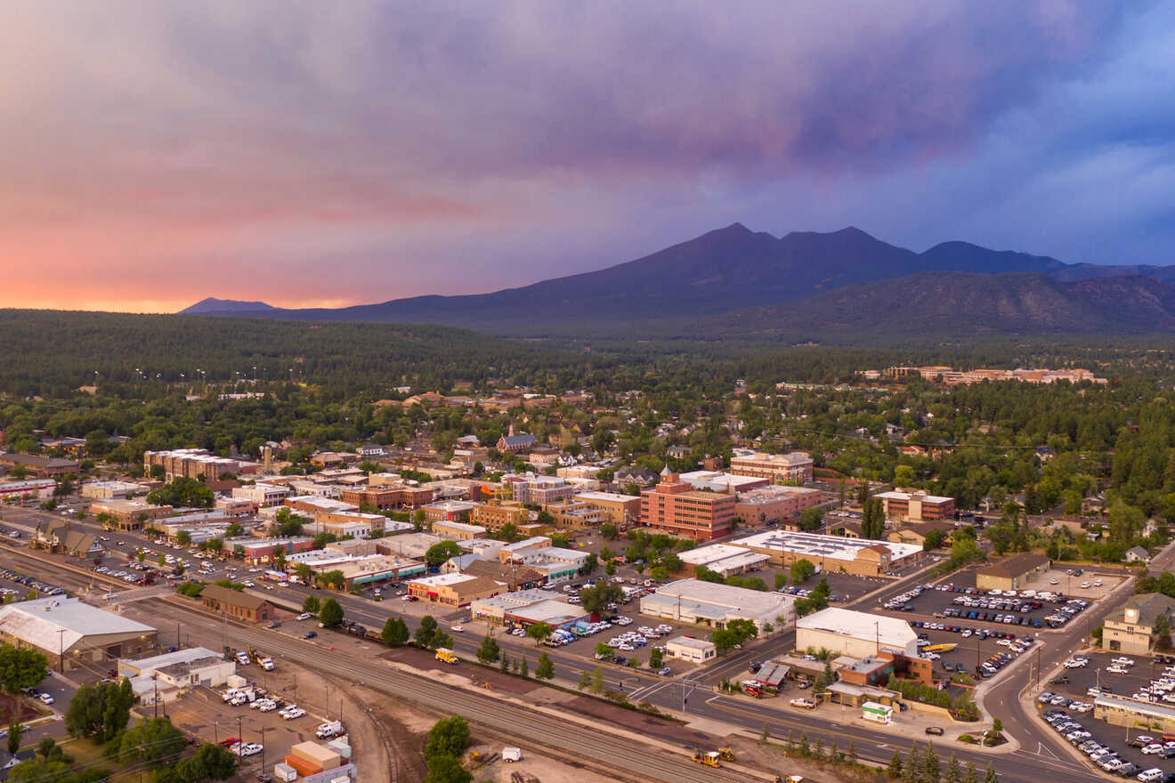 Aerial view of a town with low-rise buildings, surrounded by forest and mountains under a colorful, cloudy sky at sunset.