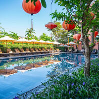 Outdoor swimming pool surrounded by lounge chairs, large umbrellas, and red lanterns hanging from trees, with plants and clear sky in the background.