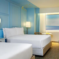 A hotel room with two neatly made twin beds, a standing lamp between them, and a window with light partially drawn curtains. The walls are painted light blue, and a small wooden chair is near the window.