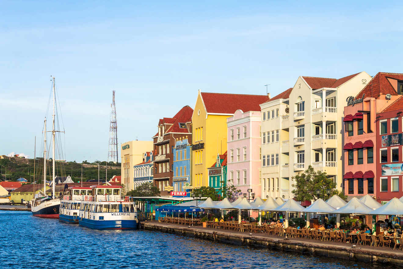 A waterfront scene with colorful Dutch colonial buildings, boats docked along the shore, and outdoor dining areas under white canopies. The water is calm under a clear sky.