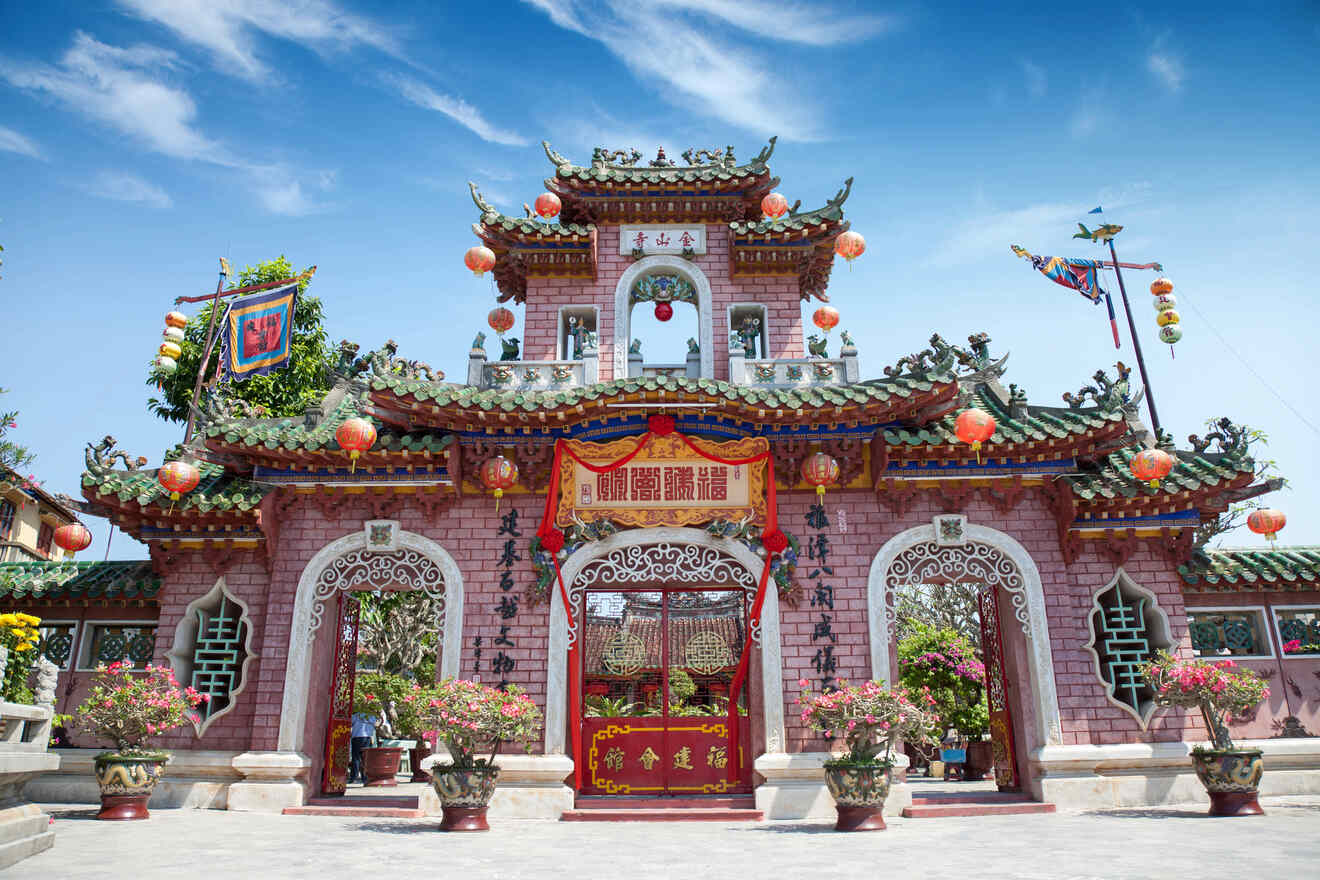 Colorful Chinese temple adorned with elaborate rooftop carvings, red lanterns, and a central archway. Flags and potted plants decorate the entrance against a clear blue sky background.