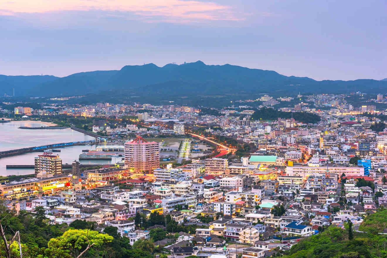 Aerial view of a coastal city at dusk with mountains in the background and numerous buildings and lights throughout.
