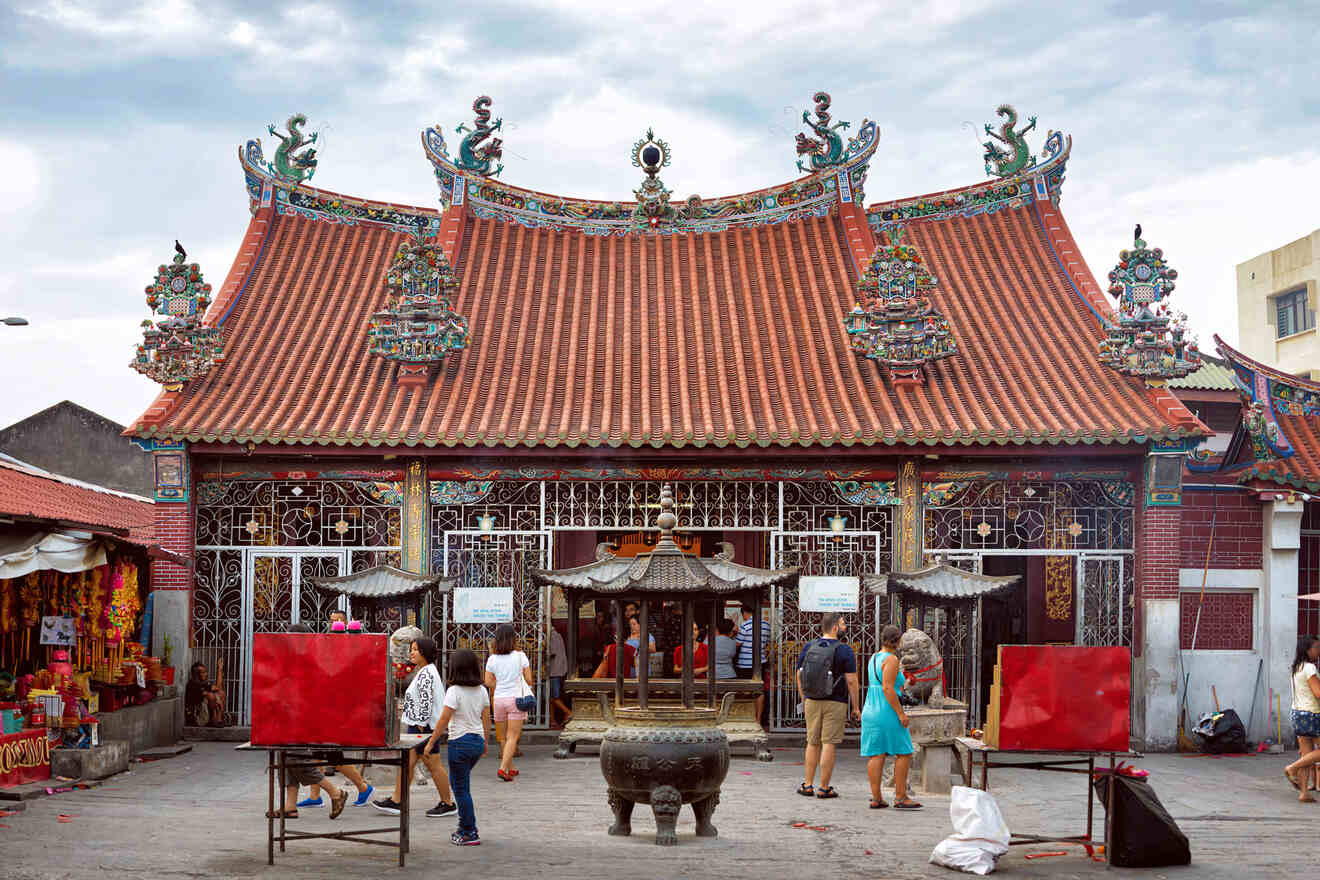 People stand and walk in front of a Malaysia temple with intricate details and ornate decorations. The temple has a red tiled roof and several small sculptures.
