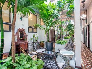 A bright indoor area with wicker chairs, tropical plants, and vintage furniture, including a wooden vanity with a mirror. Geometric patterned floor tiles and hanging lights are visible.