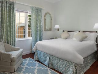 A neatly arranged bedroom features a large bed with white linens and decorative pillows, an armchair, a window with patterned curtains, a mirror on the wall, and a patterned rug on the floor.