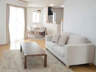 A spacious living room with a white sofa, wooden coffee table, and a beige carpet. In the background, there is a dining area with a table and chairs near windows with light curtains.