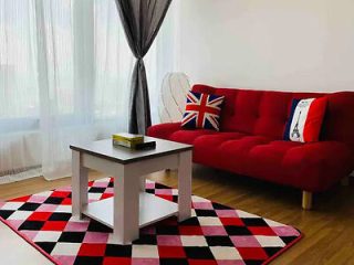 A living room with a red sofa, Union Jack cushion, black and white shelving unit, and a red, black, and white checkered rug. A window with sheer curtains and a black curtain is in the background.