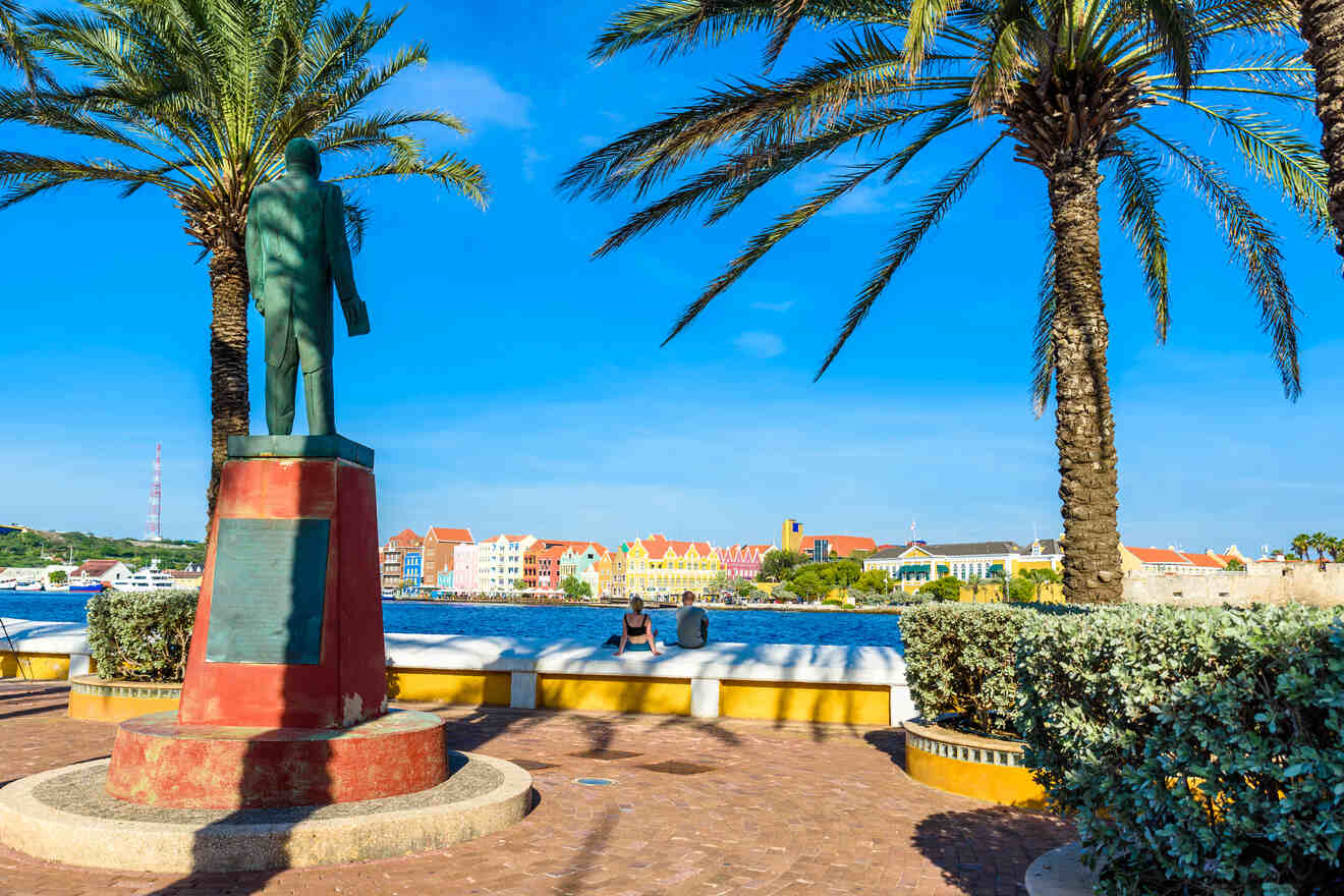 A statue on a pedestal stands between two palm trees, overlooking a waterfront with colorful buildings in the background on a clear sunny day.