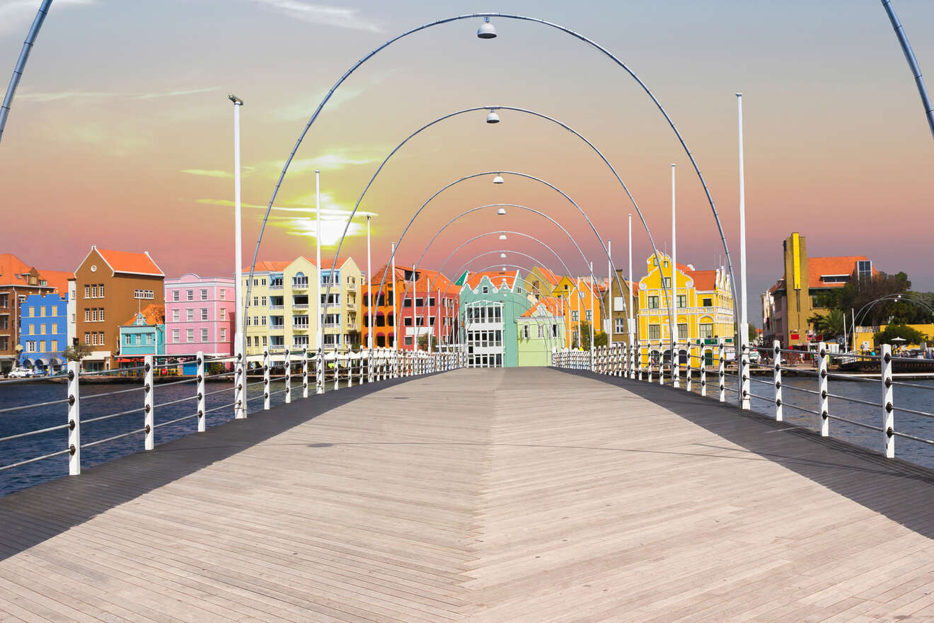 A wooden pedestrian bridge with overhead arch lights leads to a row of colorful buildings under a sunset sky.