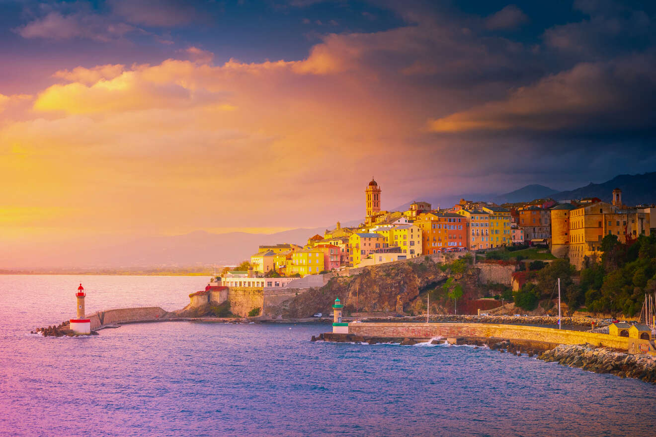 Scenic coastal town at sunset, with colorful buildings perched on a cliff, lighthouses at the edge, and dramatic skies overhead.