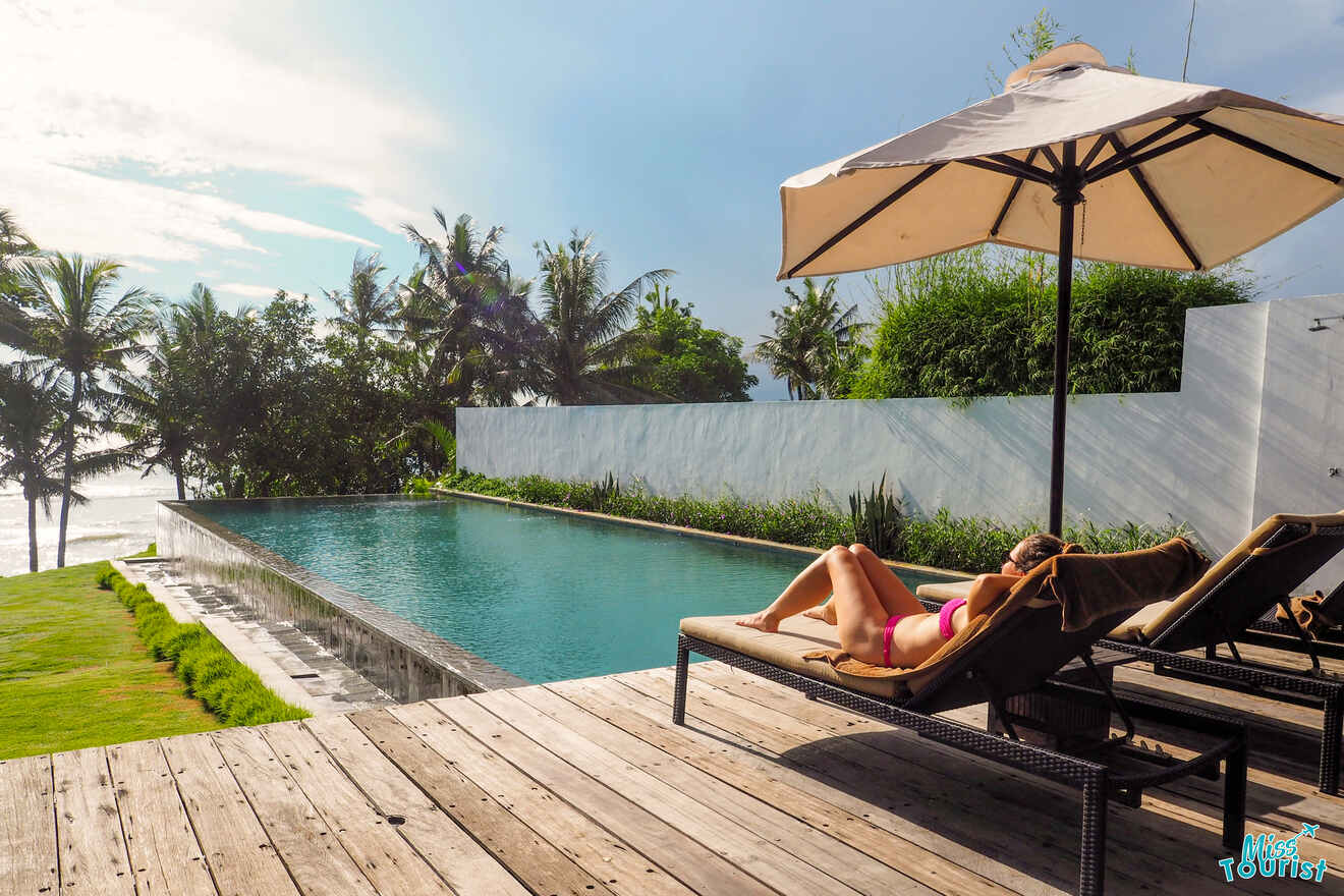 Author of the post relaxes on a sun lounger under a large umbrella beside an infinity pool, with a scenic view of palm trees and the ocean at a luxurious villa in Canggu, encapsulating a peaceful holiday retreat