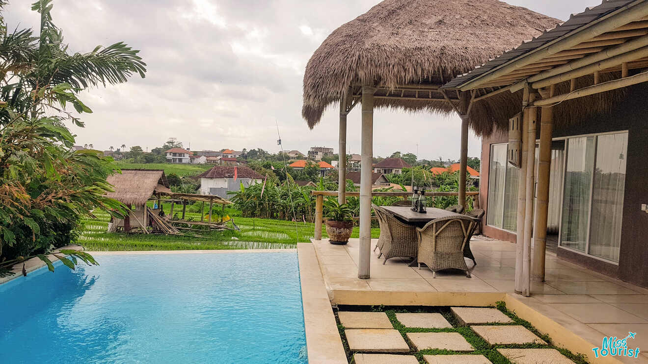A tranquil poolside view from a thatched-roof gazebo with wicker chairs overlooking a lush green rice field in Canggu, with traditional Balinese architecture in the background under a cloudy sky
