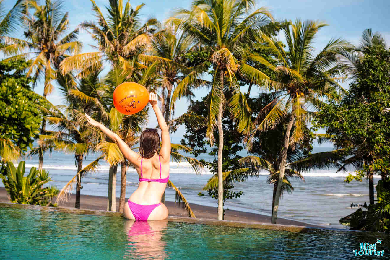 Author of the post, Yulia, in a pink bikini joyfully tosses an orange beach ball into the air while sitting on the edge of an infinity pool, with a backdrop of tall palm trees and the ocean at a beach resort in Canggu