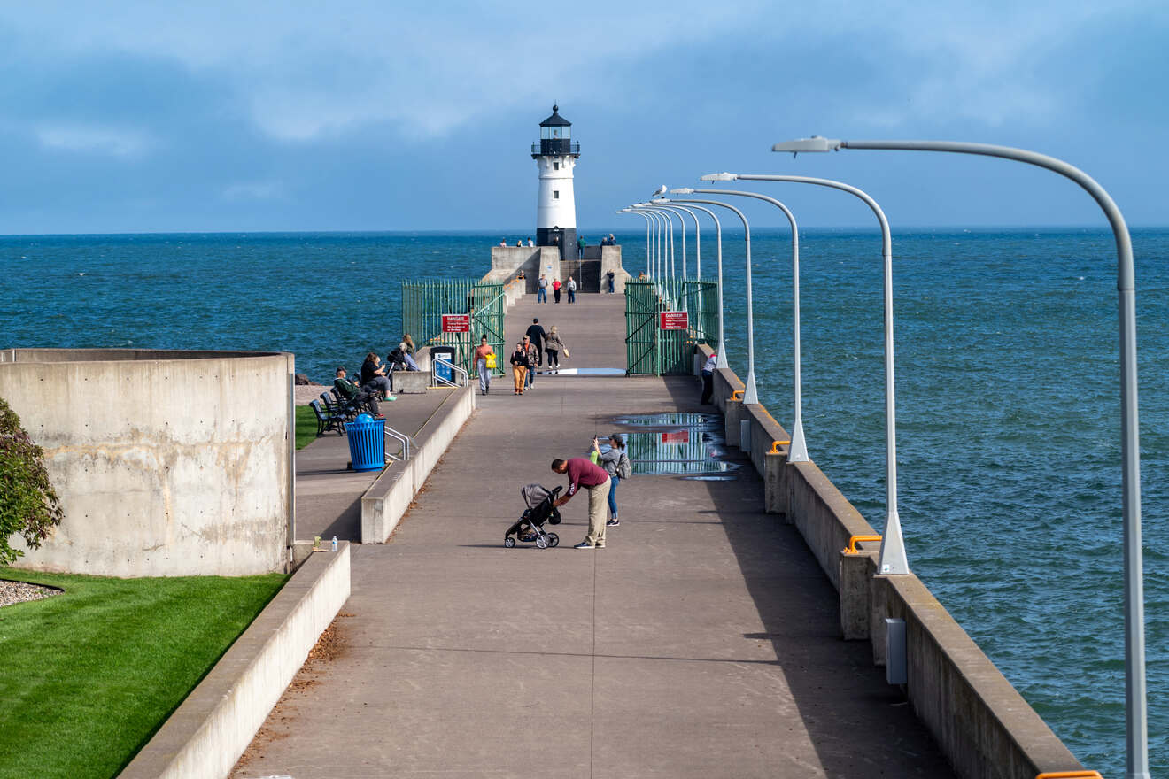 People walking on a pier leading to a lighthouse by the sea, under a partly cloudy sky, with streetlights lining the path.