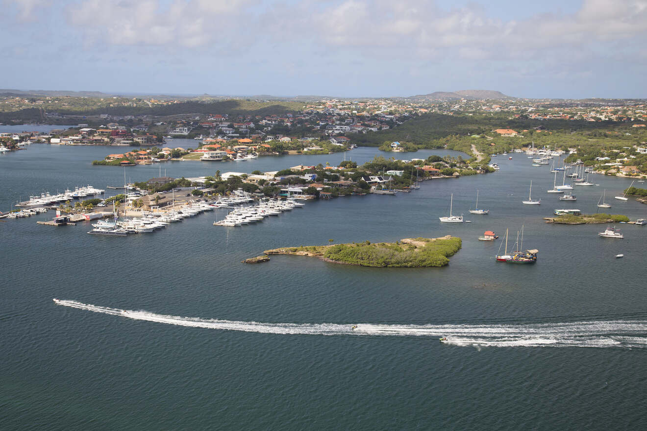 Aerial view of a coastal area featuring a marina with many boats docked, several small islands, and surrounding buildings and vegetation under a partly cloudy sky.