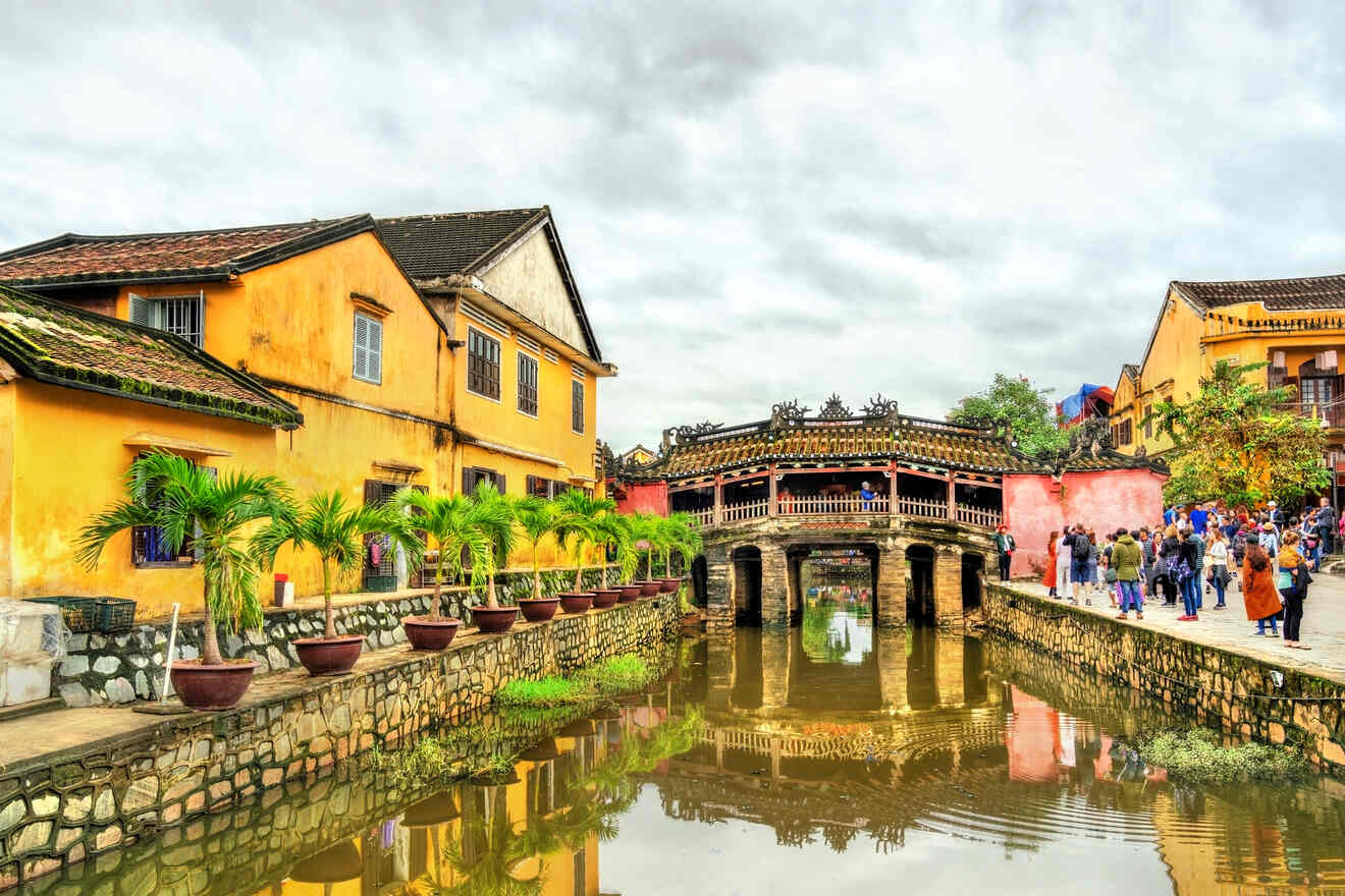 Tourists walk near the Japanese Covered Bridge in Hoi An, Vietnam, surrounded by traditional yellow buildings and a canal with a reflective surface.
