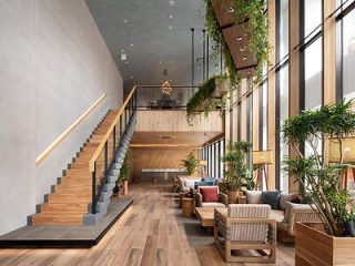 A modern, spacious lobby with wooden flooring, stairway, and furniture. Large windows let in natural light. Potted plants and hanging greenery add to the decor.