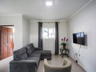 A living room with a gray sectional sofa, a beige armchair, a wall-mounted TV, a small table with flowers, and a window with dark curtains. The floor is tiled, and the walls are painted white.
