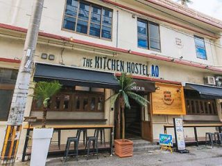 Two-story building with a sign reading "The Kitchen Hostel AO." The building has a check-in area, outdoor seating, and palm trees.