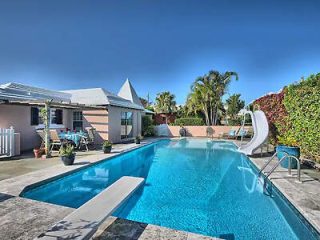 A backyard swimming pool with a slide and diving board, bordered by palm trees, shrubs, and a single-story house with patio furniture.