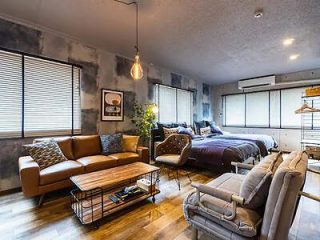 Modern studio apartment with a leather sofa, wooden coffee table, and two beds. Room includes a wall-mounted light fixture, window blinds, and eclectic decor, creating a cozy, contemporary atmosphere.