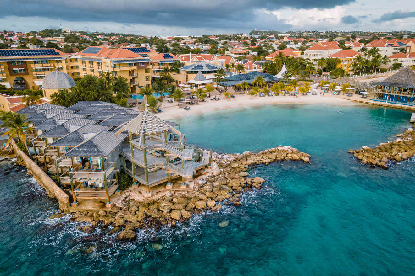 Aerial view of a coastal resort town with buildings, solar panels, and a small beach with clear turquoise water, surrounded by rocks and palm trees.