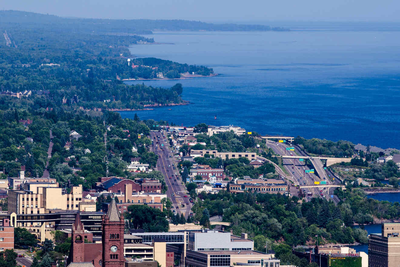 Aerial view of a coastal city with buildings, roads, and a large body of water in the background, bordered by dense green forest.