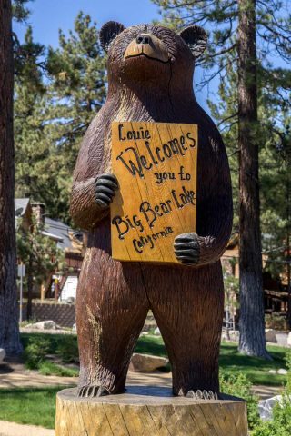 Wooden bear statue holding a sign that says "Louie Welcomes you to Big Bear Lake, California," standing among trees and buildings.