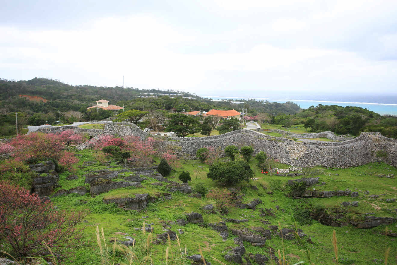 A sprawling historic stone wall winds through a lush green landscape with scattered trees and pink blooming shrubs, under a cloudy sky, with a distant view of the coastline.