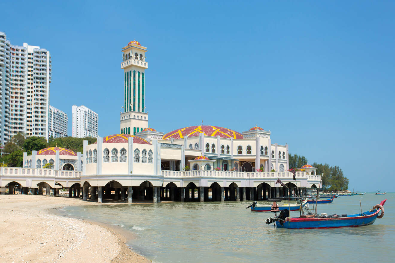 A mosque with a tall minaret stands on stilts over the water at a beach, with several boats docked nearby and modern high-rise buildings in the background.