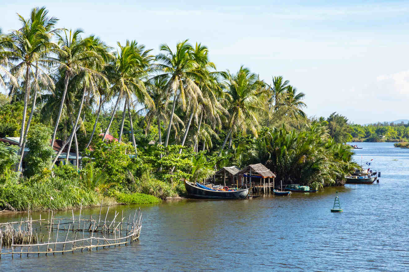 A tranquil riverside scene with palm trees, wooden huts, and boats docked at the shore. A small fishing fence is visible in the water.