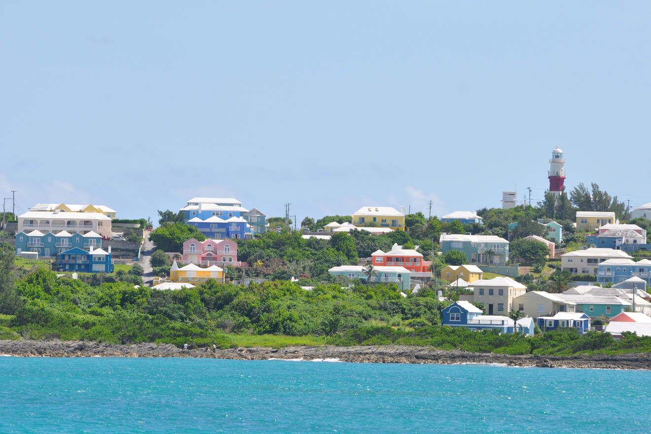 A coastal town with colorful houses on a hillside, lush greenery, and a lighthouse in the background under a clear blue sky.
