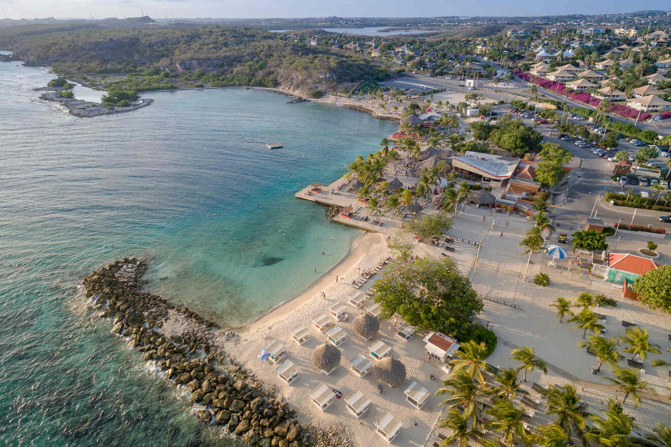 Aerial view of a beach with clear blue water, beach chairs under palapas, rocky outcroppings, and nearby buildings surrounded by greenery.