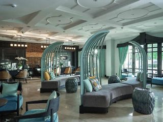 A modern, stylish hotel lobby features curved grey benches with teal accents, large windows with teal curtains, and contemporary decor under a coffered ceiling.