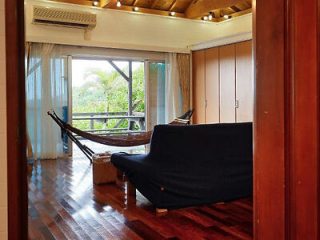 A room with wooden floors, a black couch, and a hammock near large windows leading to a balcony with greenery outside. The walls have wooden panels and there is an air conditioner above the windows.