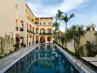 A serene outdoor pool is surrounded by a multi-story beige building with arches and balconies, along with tall palm trees and various plants.