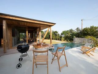 Outdoor patio with wooden chairs, a grill, and a table next to a swimming pool. The patio is attached to a wooden house, surrounded by greenery under a clear sky.