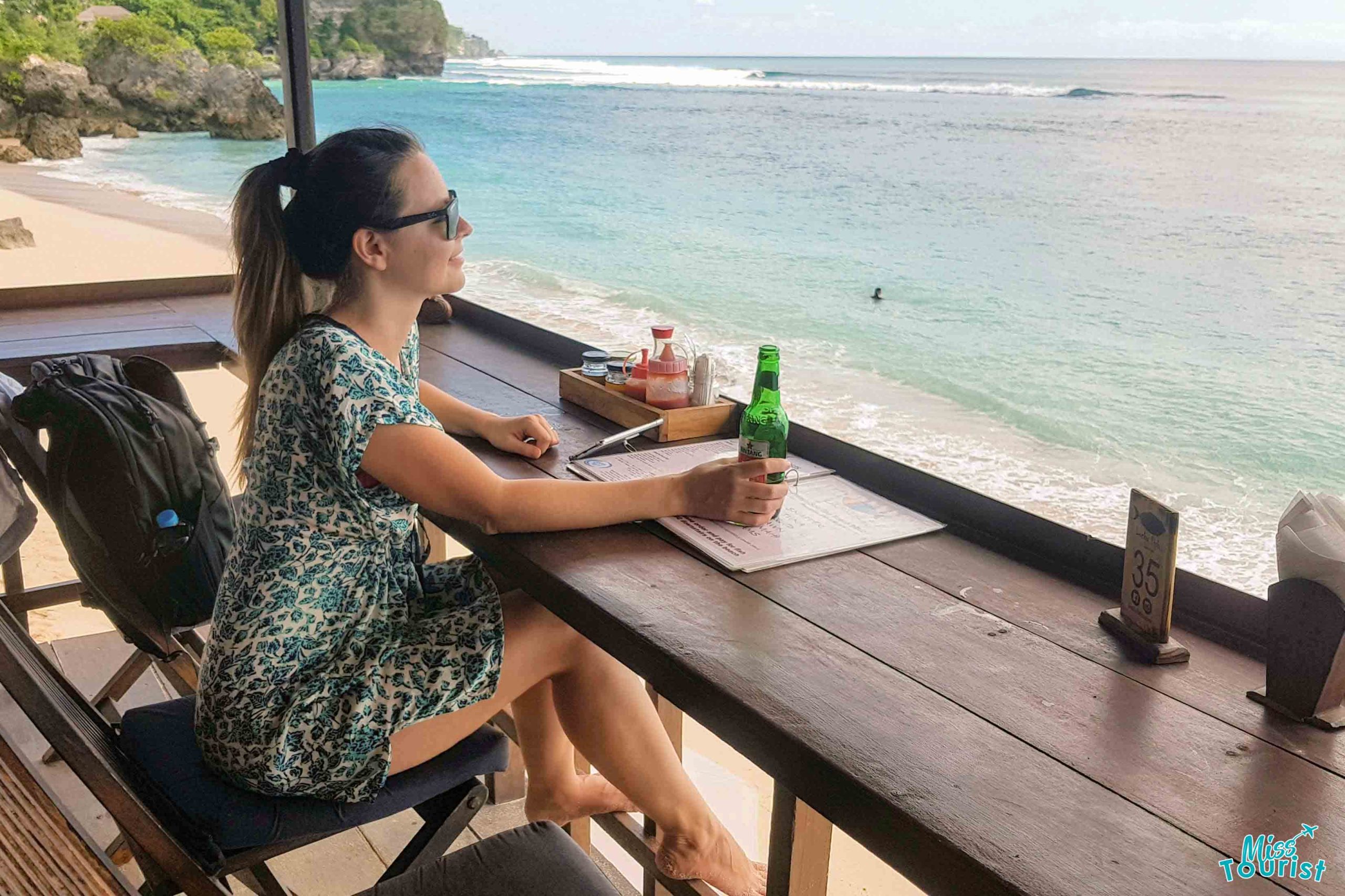 Yulia, author of the post, in a blue printed dress and sunglasses sits at a beachside bar, gazing out at the clear turquoise sea and surfers riding waves, with a beer and menu on the table, embodying a relaxed dining experience in Canggu