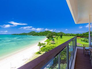 A balcony overlooks a clear turquoise sea, a sandy beach, and a grassy area with trees, all under a bright blue sky.