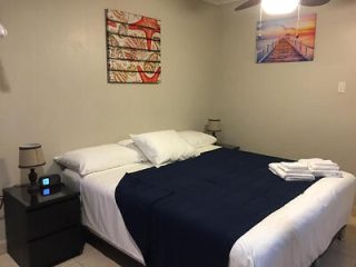 A neatly made bed with blue and white linens in a small bedroom. Two bedside tables with lamps are on either side, and framed artwork hangs on the wall. A stack of folded towels is on the bed.