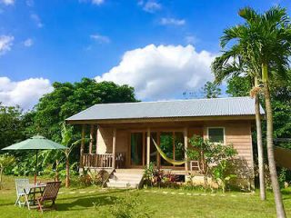 A small wooden house with a metal roof set in a grassy yard with a table, chairs, and green umbrella. A hammock hangs on the front porch. Surrounded by tropical trees under a blue sky with white clouds.