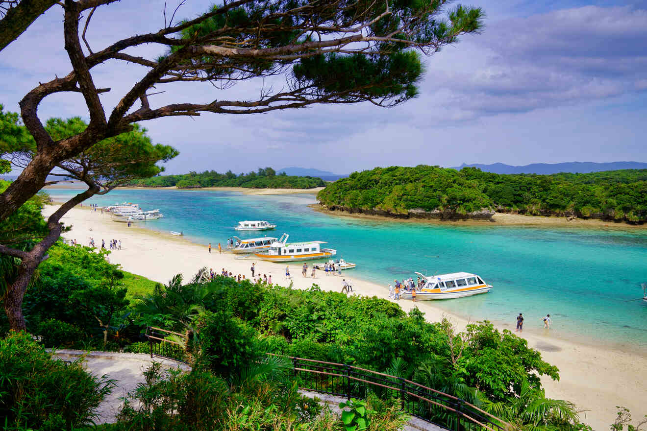 People walking on a sandy beach near turquoise water with boats anchored close to shore. The area is surrounded by lush greenery and trees with mountains in the distance under a partly cloudy sky.