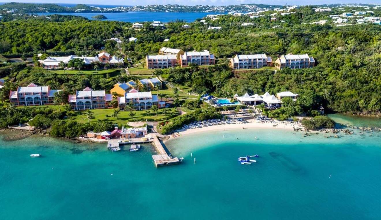 Aerial view of a coastal resort with buildings, a beach, dock, boats, and lush greenery, overlooking a body of water with distant land and water features.