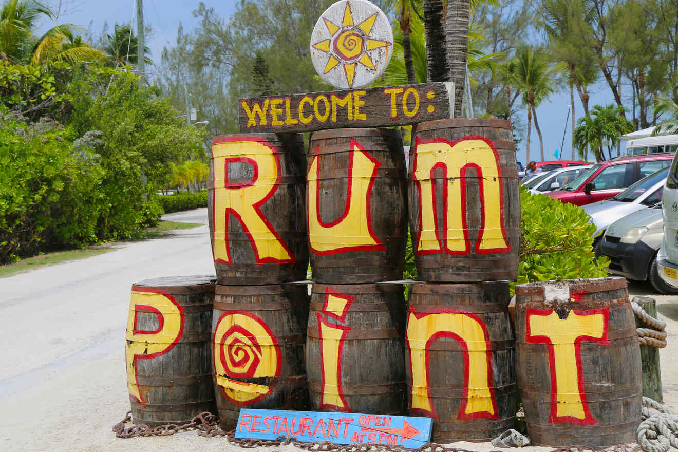 Sign at Rum Point made from stacked barrels with painted letters, a sun design, and a smaller sign at the bottom advertising an open restaurant. Trees and parked cars visible in the background.