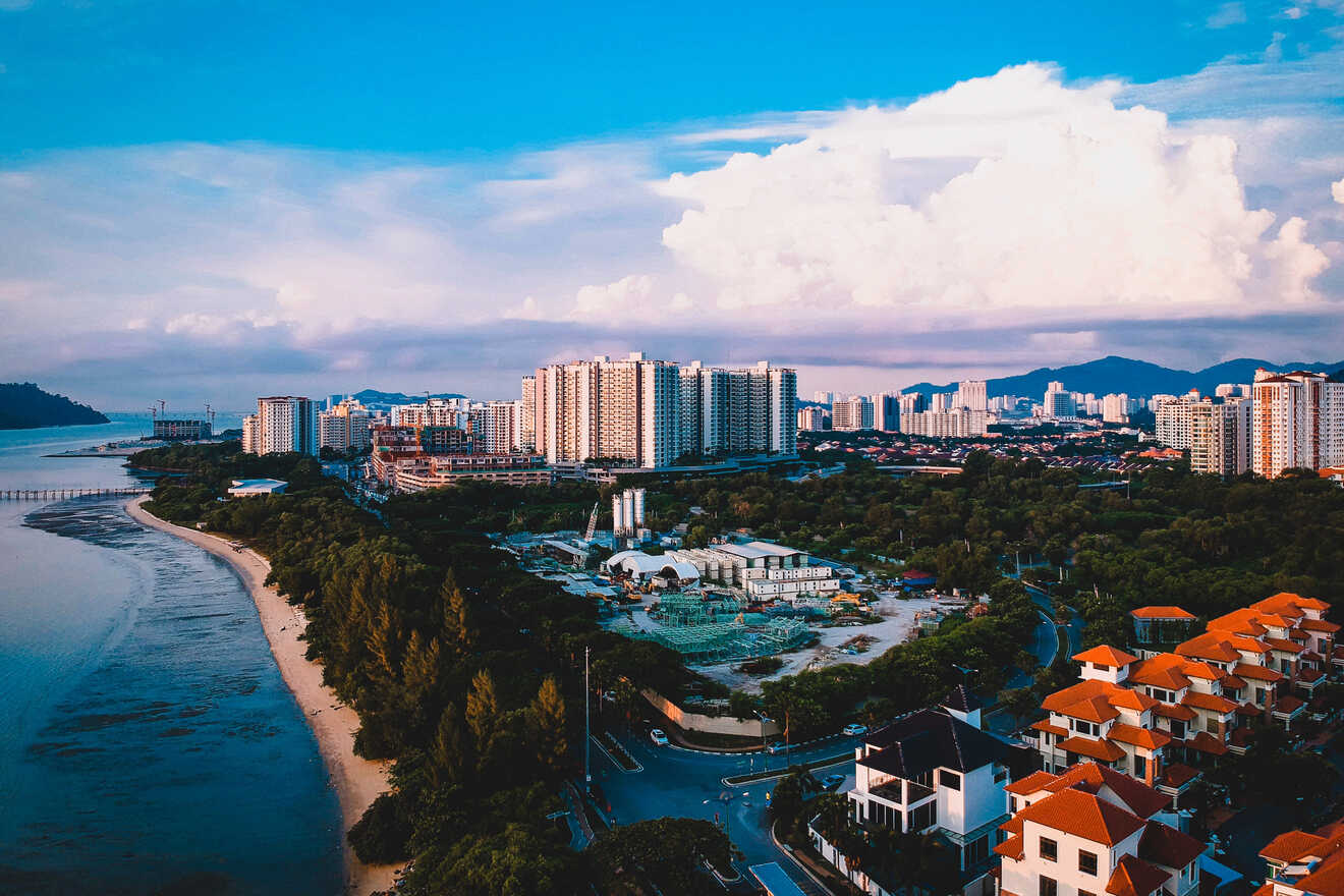 Aerial view of a coastal city with high-rise buildings, a construction site, lush greenery, a beach, and a body of water under a blue sky with clouds.