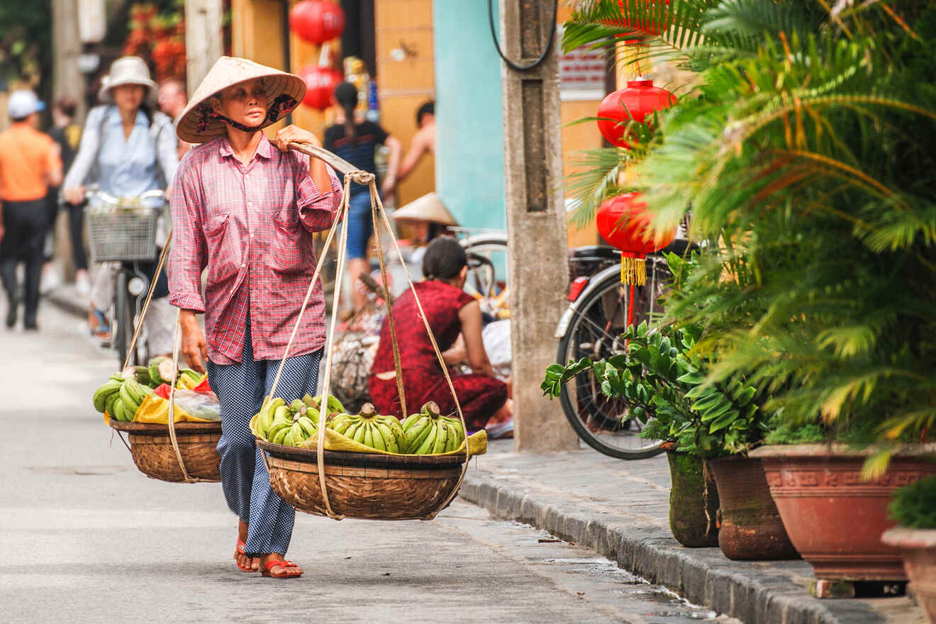 A person wearing a conical hat carries a traditional bamboo yoke with baskets full of bananas on a street with red lanterns and greenery.