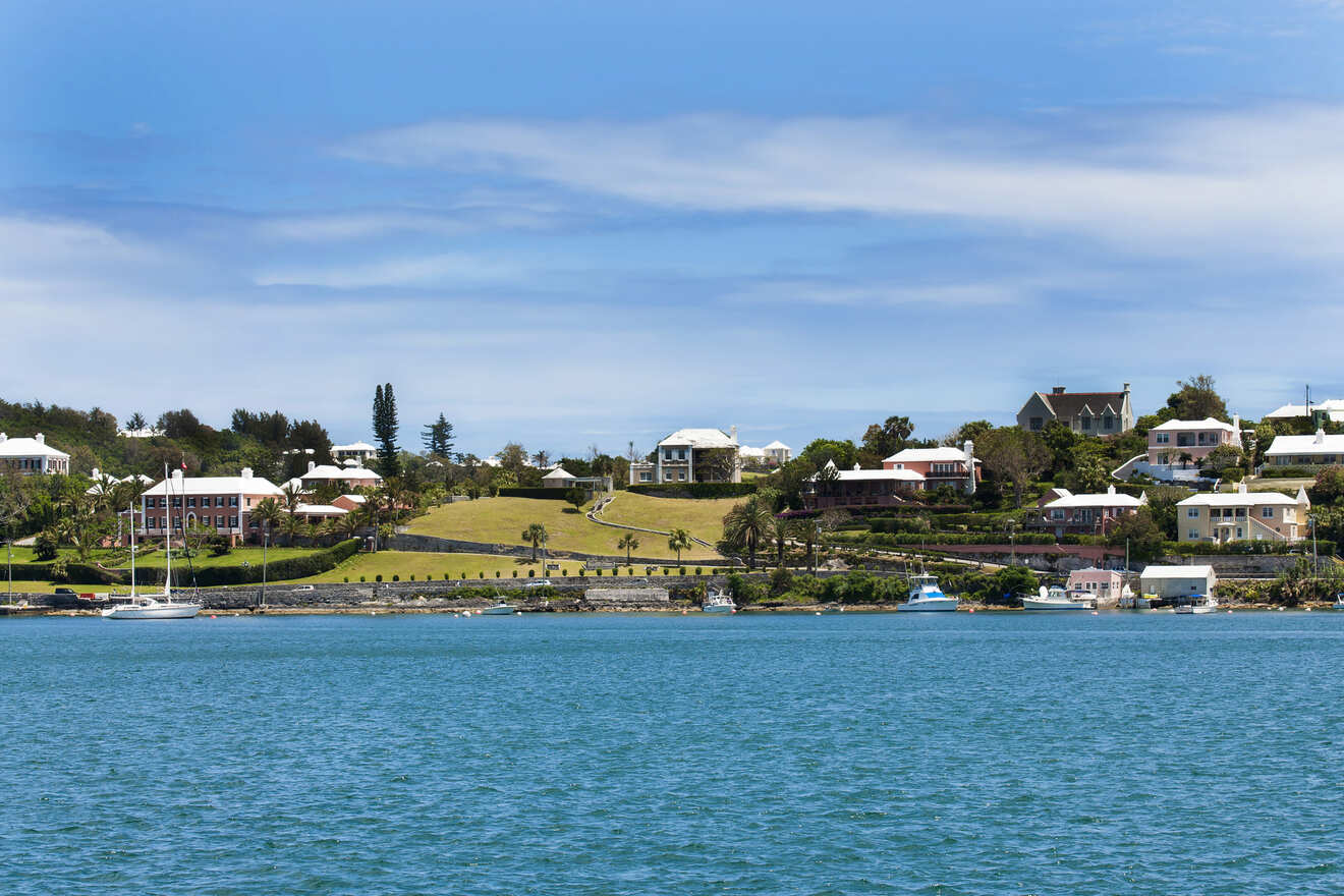 A coastal view featuring lush green hills, residential homes, and a few docked boats, all under a blue sky with scattered clouds.