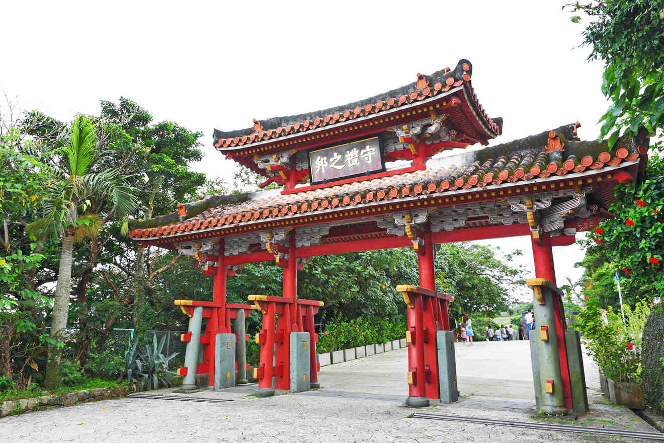 A traditional red wooden gate with ornate roof tiles stands at the entrance to a path, surrounded by trees and greenery. People are visible in the background walking through the gate.